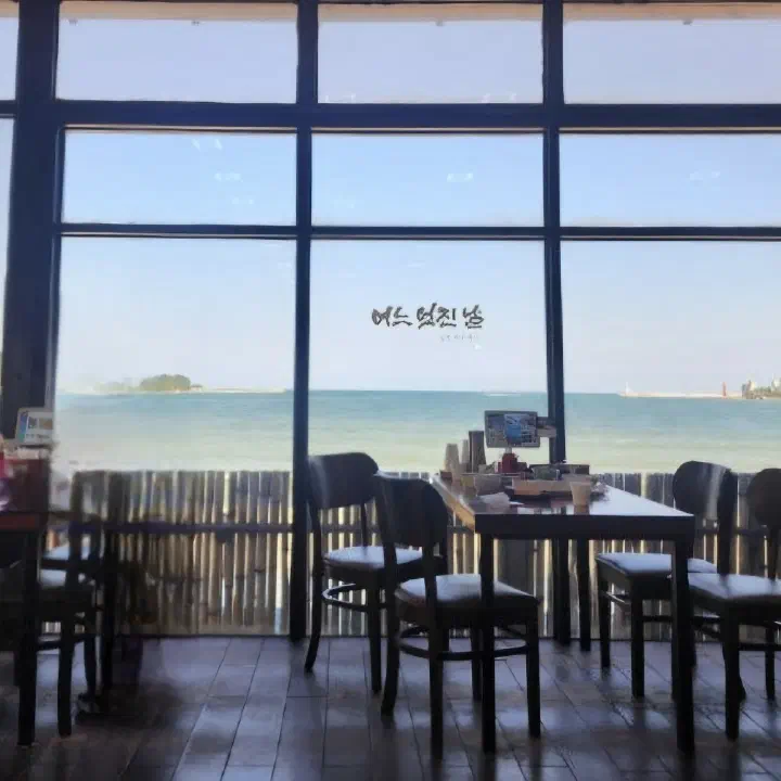 어느멋진날 카페 통창 너머로 바다와 섬이 펼쳐지고 창가에 나무 테이블과 의자가 놓여 있다