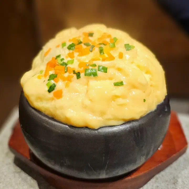 검은 돌그릇에 부드럽게 부풀어 오른 노란 치즈 계란찜이 담겨 나온 모습을 가까이서 찍은 사진