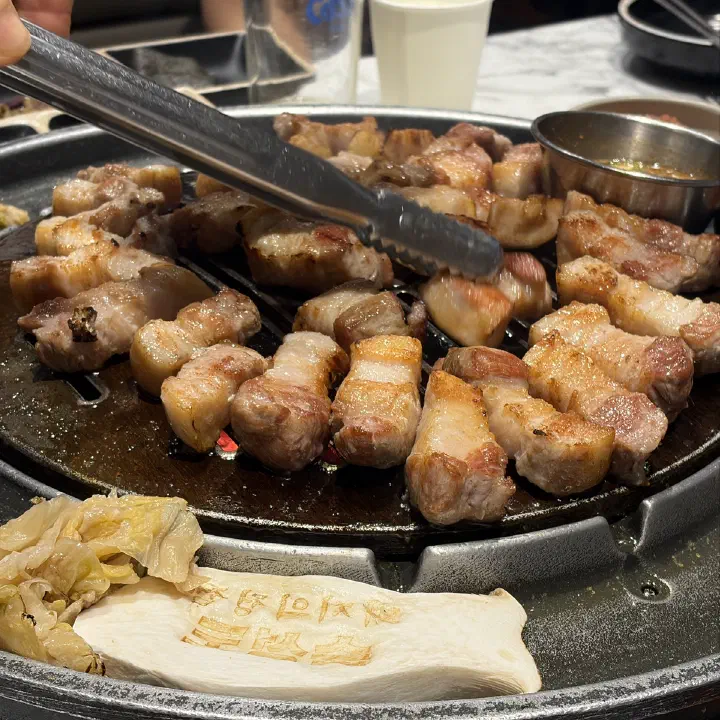 숙성도 중문점 불판 위에서 집게로 뒤집는 중인 노릇하게 구워진 삼겹살 한입 크기 고기 클로즈업