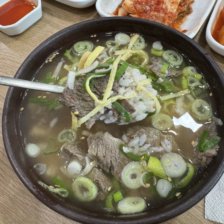 어두운 뚝배기에 담긴 맑은 곰탕에 얇은 소고기 조각과 파, 채 썬 노른자 지단이 올라가 있고, 숟가락으로 밥을 떠 올린 모습